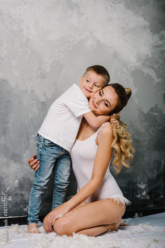 Smiling mom with son sitting on the floor. Fashion models plays with feathers. Happy family portrait in casual style clothes. Nice family wearing jeans isolated on grey wall. © Serhii