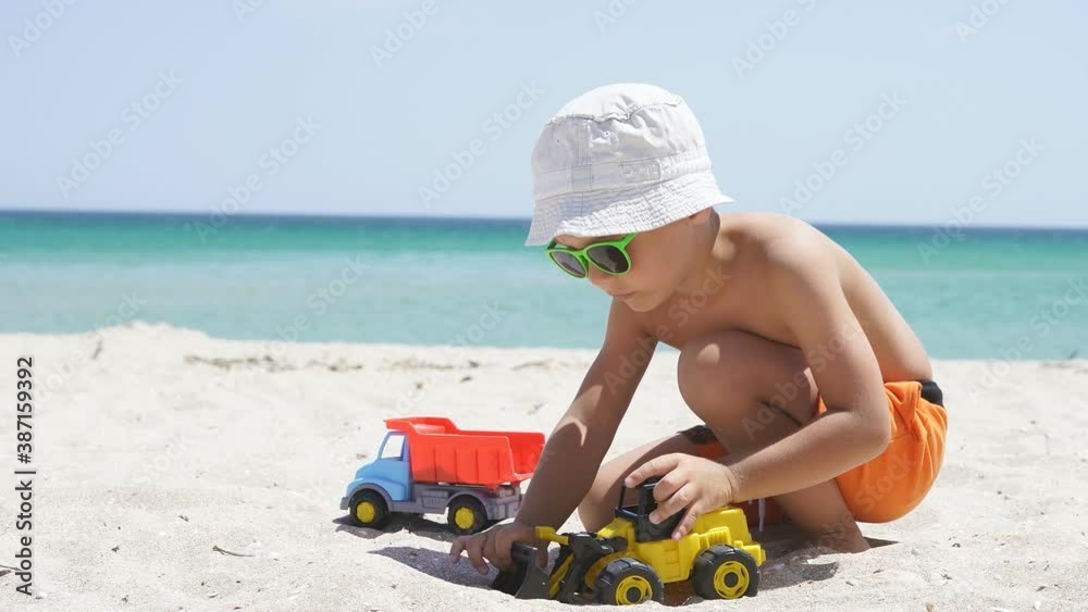 boy dressed in shorts, a Panama hat and sunglasses plays with cars on a sandy beach, against the background of a beautiful azure sea and sky on a Sunny day. boy dressed in shorts, a Panama hat and sunglasses plays with cars on a sandy beach, against the background of a beautiful azure sea and sky on a Sunny day.