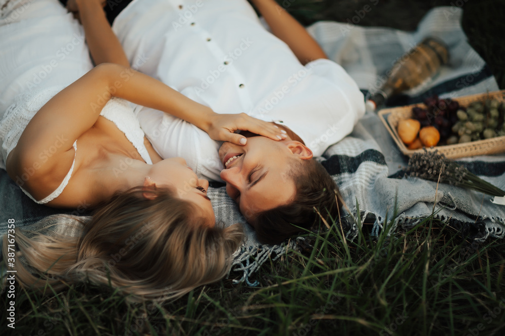 Happy young couple in the park is relaxing at the summer picnic. They lying on blanket on green grass, looking each other and smiling.