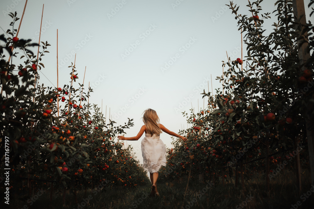 Pretty young girl in white dress jumping on green grass in the apples orchard. Back view.