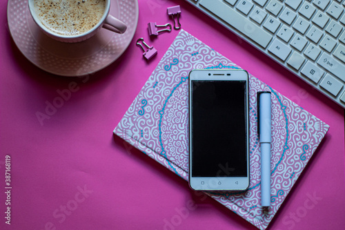 Creative flat lay photo workspace: keyboard, coffee, mobile, notebook, pen and some paper clips, pink background, selective focus, free copy space