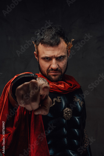 Obraz na plátně Serious and bearded roman emperor dressed in dark armour and red cloak points finger at camera in dark background