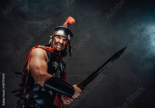 Canvas Print Armoured savage and brutal roman warrior in red cloak with helmet attacks holding sword in smokey background