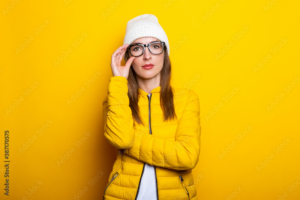 Pensive young woman holding glasses in yellow jacket on yellow background.
