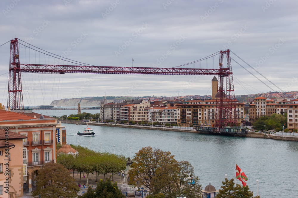 Fototapeta premium Portugalete, Spain - 10/17/2019: Biscay bridge flying with gondola and boat on river Nervion, toned. Portugalete landmark. Famous bridge called Puente de Vizcaya near Bilbao. Riverbank of Portugalete.