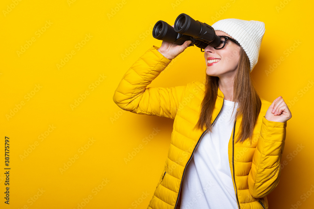 Young woman in a yellow jacket smiling looks through binoculars on a yellow background. Young woman in a yellow jacket smiling looks through binoculars on a yellow background.