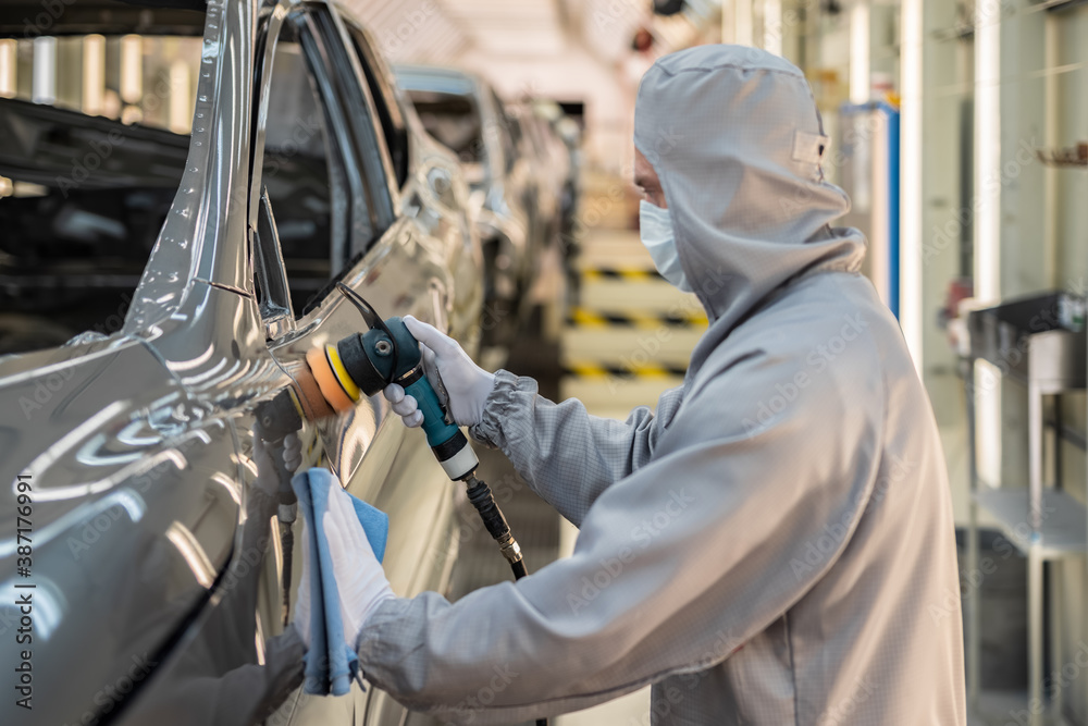 An employee of the car body paint shop with a medical mask on his face polishes the painted surface