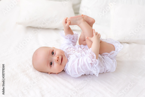 a small baby girl 6 months old is lying on a light bed in a beautiful white bodysuit and smiling at the camera, the baby is lying on her back and holding her legs