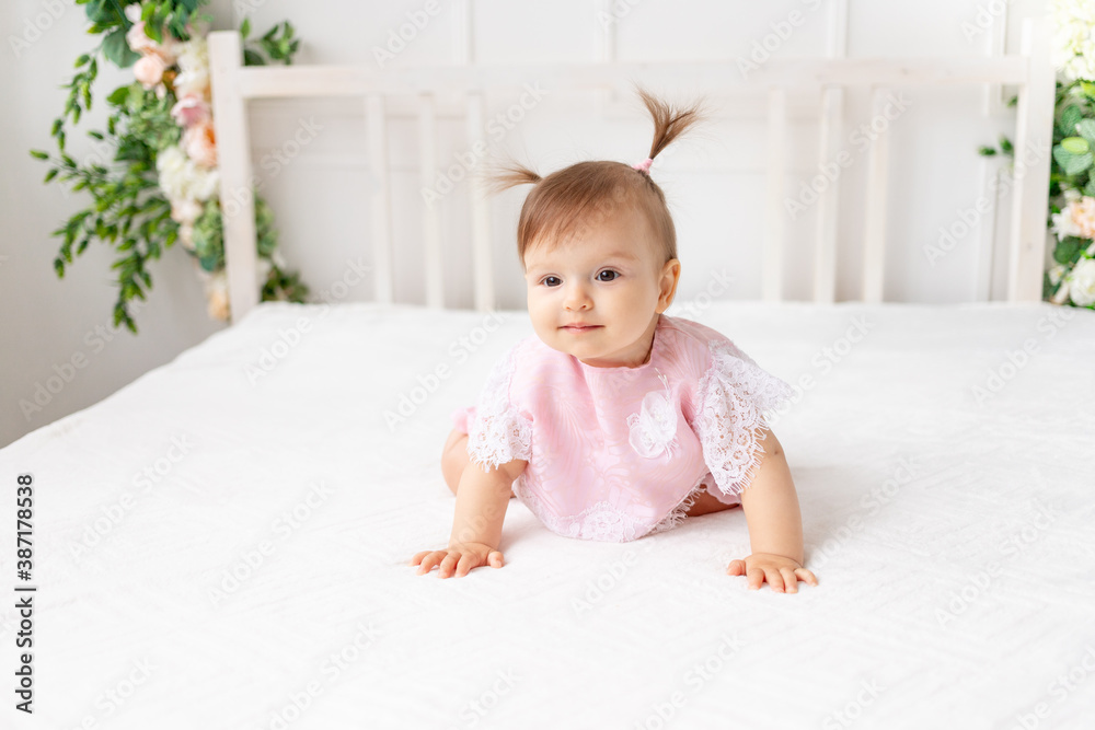cheerful baby girl six months old sitting in a bright beautiful room on a white bed in pink clothes and smiling cheerful baby girl six months old sitting in a bright beautiful room on a white bed in pink clothes and smiling