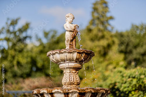Closeup of the top of an old fountain with a sculpture of a child with flowing water with lush green trees and a blue sky in a blurred background, sunny summer day © Emile