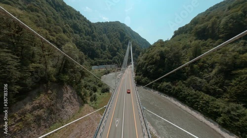 Fpv sport drone reelsteady shot. Red cabriolet car drives along contemporary cable-stayed bridge over Mzymta river curving between mountains in Sochi area first point drone view photo