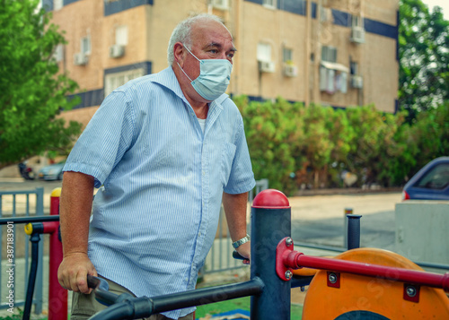 An elderly man exercises with an outdoor machine near his residential building. He wears a face mask during the pandemic. He has a red face. Because of his muscles warm-up for the workout ahead.
