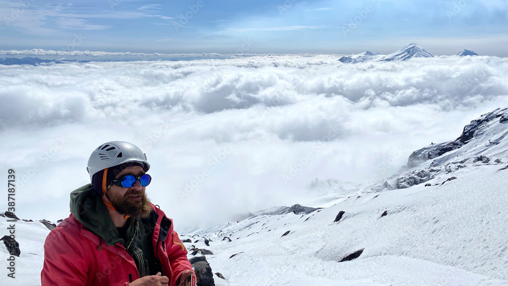 A climber in a helmet and sunglasses stands above the clouds on his way to the top of the mountain.