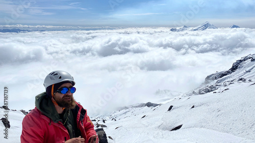 A climber in a helmet and sunglasses stands above the clouds on his way to the top of the mountain.