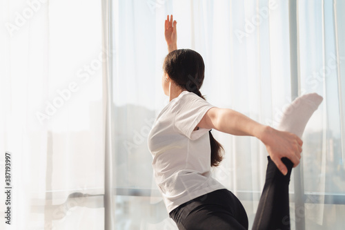 A young woman in sportswear is stretching in the morning in the bedroom. Windows in the background. Yoga at home. View from the back. Copy space