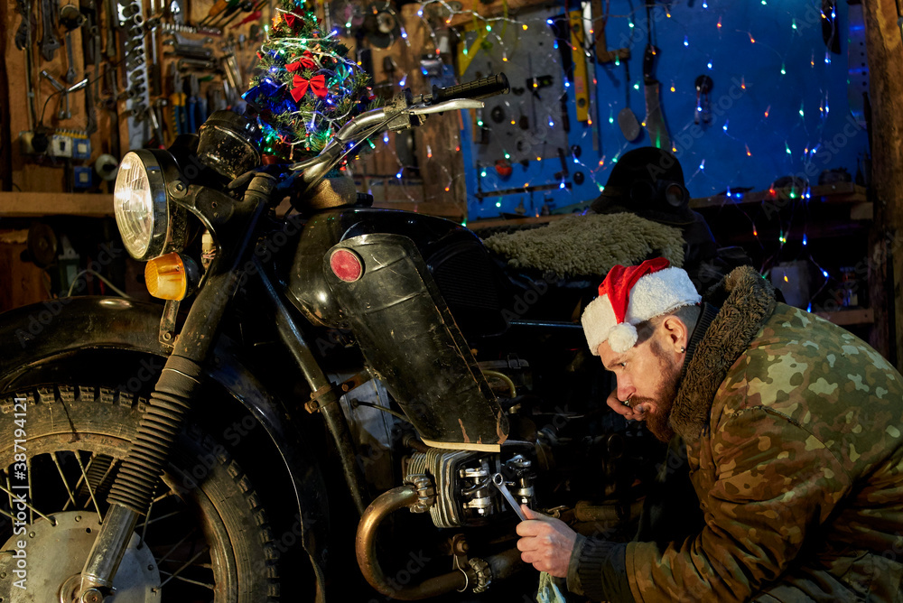 A man in a santa claus hat repairs a motorcycle in the garage on New Year's Eve.