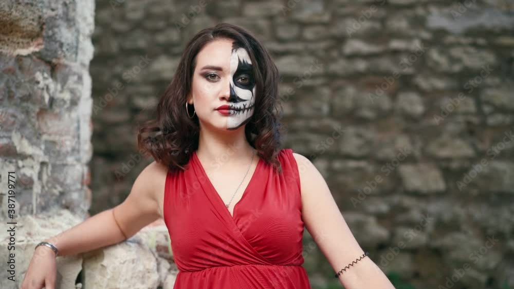 Portrait beautiful woman in a red evening dress and awesome Halloween makeup stands against the backdrop of an abandoned building. Greasepaint for the Holiday on October 31. Slow motion Portrait beautiful woman in a red evening dress and awesome Halloween makeup stands against the backdrop of an abandoned building. Greasepaint for the Holiday on October 31. Slow motion
