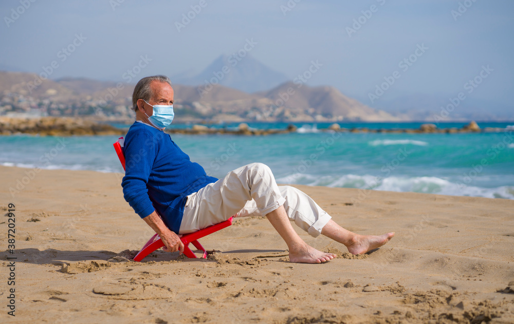 Senior pensioner relaxed in beach during new normal - retired old man on his 70s wearing surgical mask against covid19 sitting at the sea thoughtful and contemplative