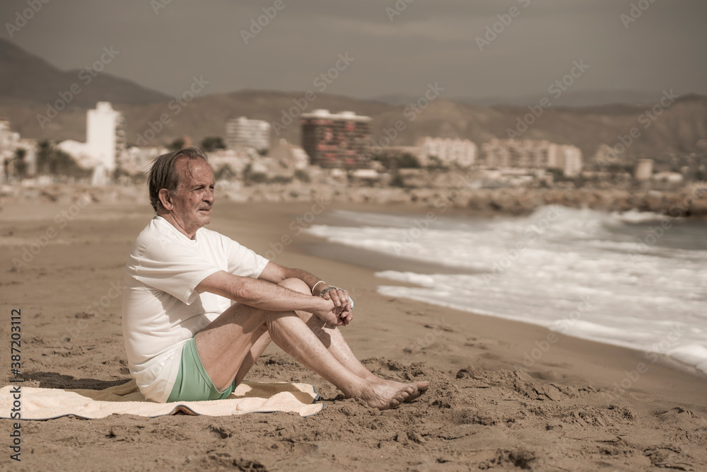 Senior pensioner sitting relaxed on the beach - retired old man on his 70s looking at the sea thoughtful and contemplative in health concept