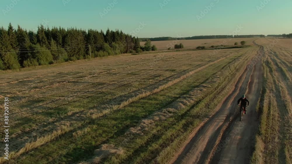 Man in green helmet rides enduro electro full suspension mountain bicycle e-bike along rural dirty road dust from the wheels past field and wood on sunny autumn evening aerial view. Man in green helmet rides enduro electro full suspension mountain bicycle e-bike along rural dirty road dust from the wheels past field and wood on sunny autumn evening aerial view.