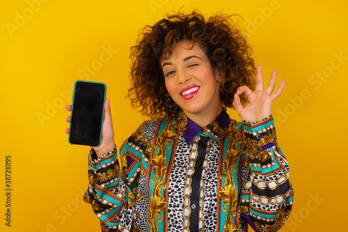 Excited woman showing smartohone blank screen, blinking eye and doing ok sign with hand. Studio shot of shocked girl holding smartphone with blank screen. Advertisement concept. photo