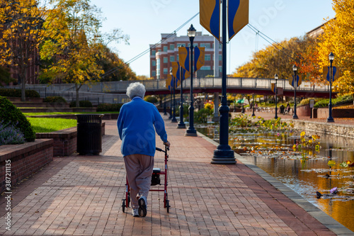 An elderly caucasian woman wearing blue sweater, track pants and sneakers is slowly walking in parks with a rollator walker. It is a sunny autumn day in Frederick, Maryland with river on one side photo
