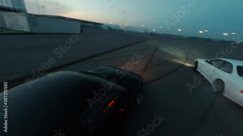 Sports automobiles drive fast drifting smoke from wheels on empty plain road in contemporary night city first point view fpv sport drone follow close shot, round evening sunset panorama photo