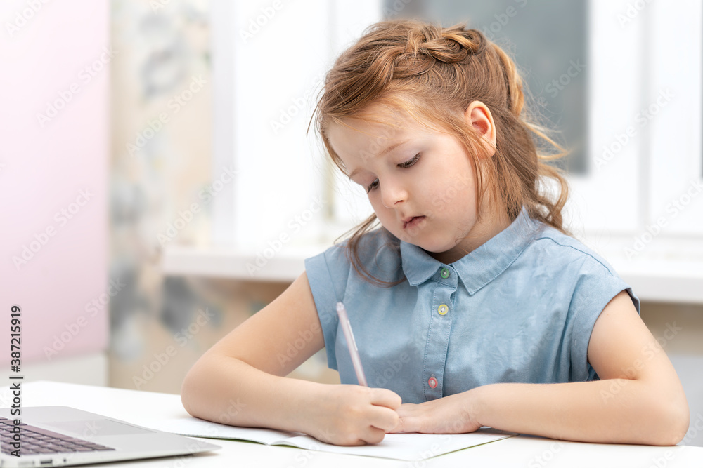 smart beautiful girl with long wavy hair sits at a white table and writes in a notebook in front of a laptop, does homework. Distance learning at home, a new reality