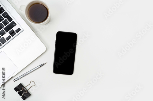 a phone with a screen mockup, a coffee cup of writing supplies, a pens, a notepad on a white wooden table background. Top view of the work area, copy space