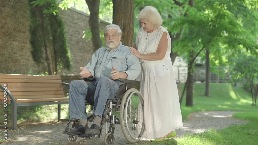 Old man sitting in wheelchair and talking to senior beautiful woman. Portrait of Caucasian disabled man spending sunny day outdoors with loving wife. Unity and support concept. Old man sitting in wheelchair and talking to senior beautiful woman. Portrait of Caucasian disabled man spending sunny day outdoors with loving wife. Unity and support concept.
