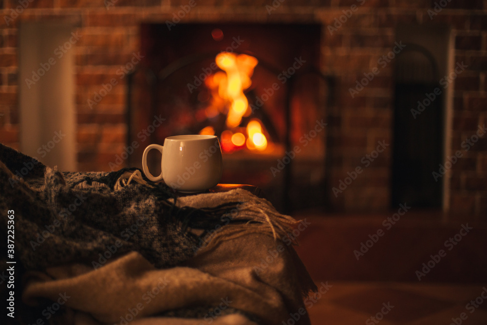 Mug with hot tea standing on a chair with woolen blanket in a cozy living room with fireplace. - obrazy, fototapety, plakaty 