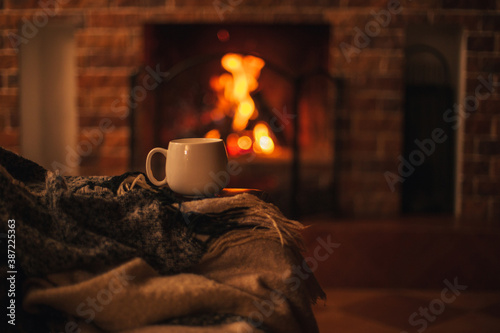 Mug with hot tea standing on a chair with woolen blanket in a cozy living room with fireplace. photo