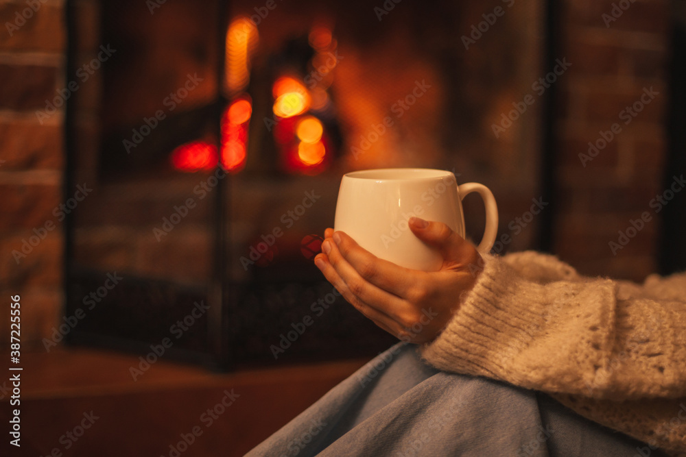 Young woman wearing white woolen knitted sweater enjoying hot tea near fireplace in a cozy living room. - obrazy, fototapety, plakaty 