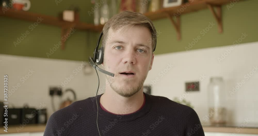 Male teacher using headset during video call and looking at camera, working from home at kitchen table during pandemic