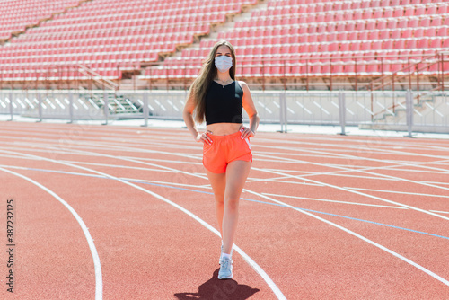 Young fit female in sportswear and protective mask for coronavirus on track during outdoor workout