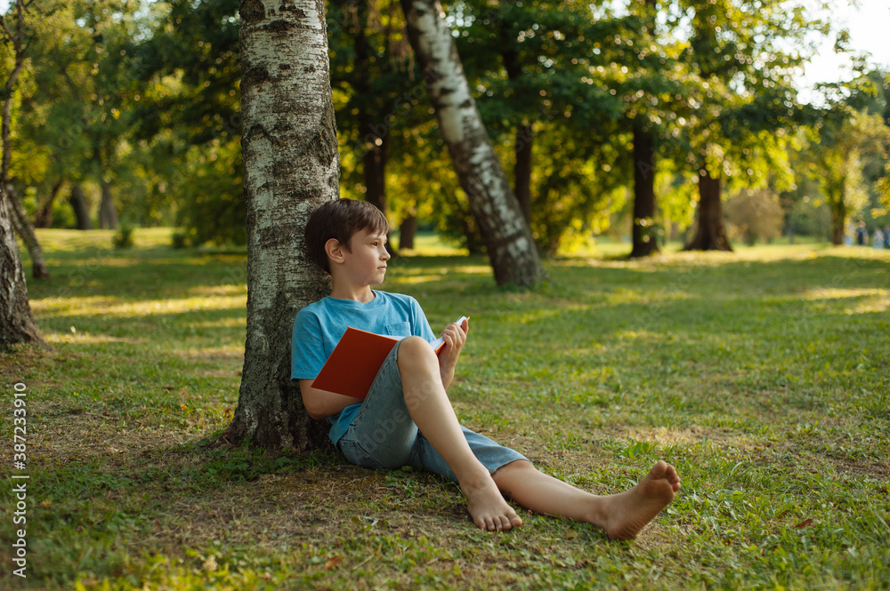 A child holding his journal and looking away leaning on a tree in park