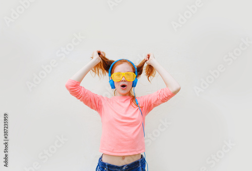 a teenage girl with red hair wearing yellow glasses and blue headphones is dancing near a white wall