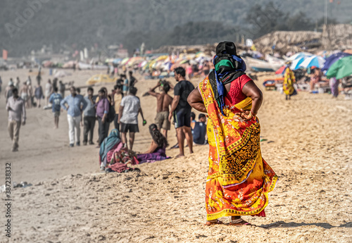 Unidentified beautiful indian women in colorful national clothes stands on beach in powerful pose and looking forward at beach full of walking tourists and local people, view from women back side