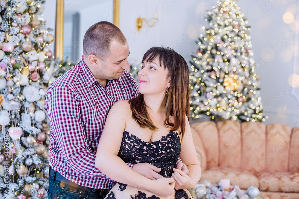 Middle-aged couple in elegant clothes on a background of Christmas Interior: snow covered trees, white fireplace, luxury furniture. Young woman in a lush, evening dress among garlands of lights Middle-aged couple in elegant clothes on a background of Christmas Interior: snow covered trees, white fireplace, luxury furniture. Young woman in a lush, evening dress among garlands of lights