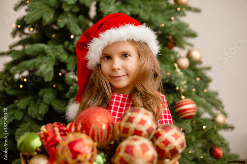 cute little girl in a santa hat holds a basket of toys against the background of the Christmas tree. Children dresses up christmas tree for new year