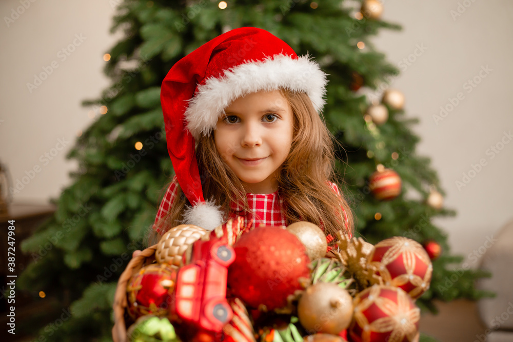 cute little girl in a santa hat holds a basket of toys against the background of the Christmas tree. Children dresses up christmas tree for new year