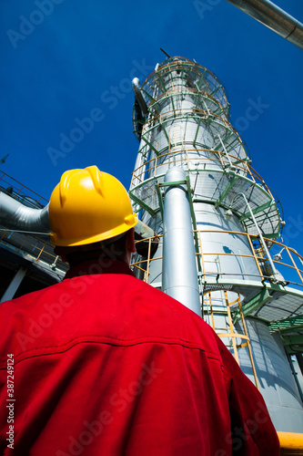 Refinery worker in red work wear and yellow hardhat on oil refinery and gas processing plant. Looking on Distillation column on blue sky background.