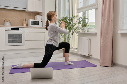 A blonde woman doing yoga stands in a warrior pose. Training in sports and yoga online. There's a laptop on the floor.