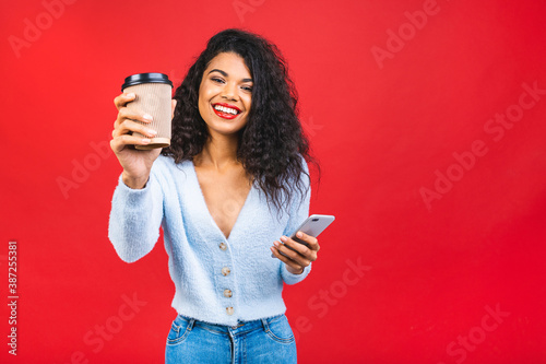 Happy young african american woman casually dressed standing isolated over pink background, holding mobile phone and cup of coffee or tea. © denis_vermenko