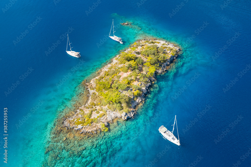 aerial view to little island in emerald water between three sailing yachts