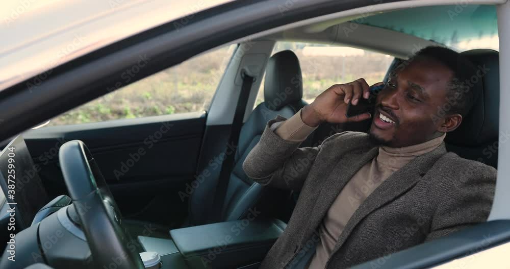 Close up of young African American man traveling in electric car and calling on smartphone. Handsome happy male speaking on mobile phone while sitting in modern vehicle. Automobile concept Close up of young African American man traveling in electric car and calling on smartphone. Handsome happy male speaking on mobile phone while sitting in modern vehicle. Automobile concept