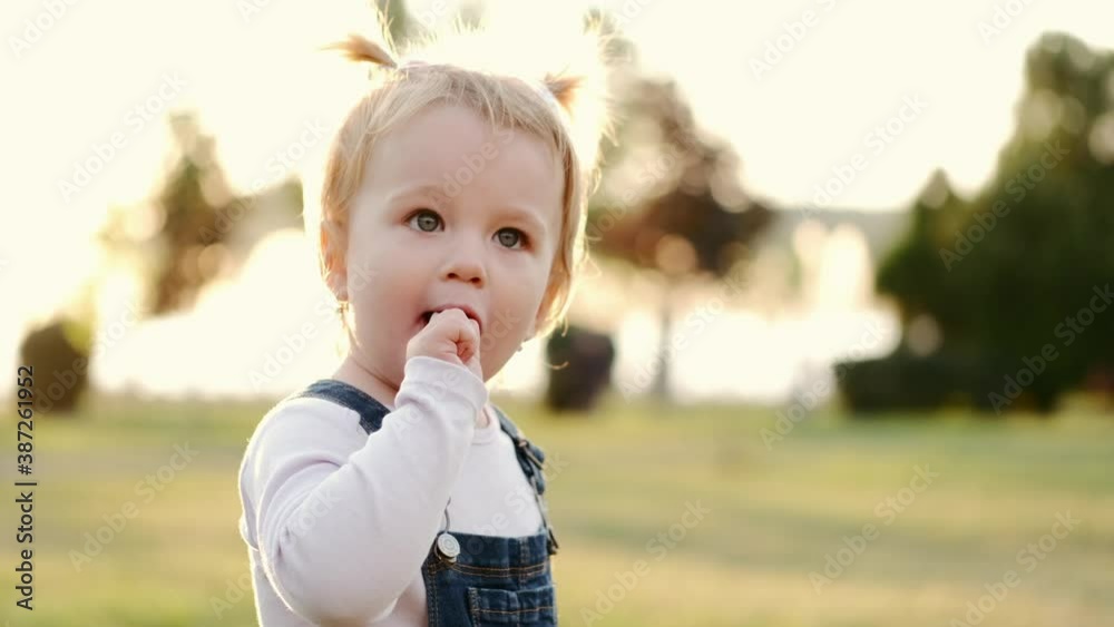 Little girl sucks chupa chups outside in the park. Close-up funny baby with two tails. A girl holding a candy in her hands, takes it in her mouth and licks Little girl sucks chupa chups outside in the park. Close-up funny baby with two tails. A girl holding a candy in her hands, takes it in her mouth and licks