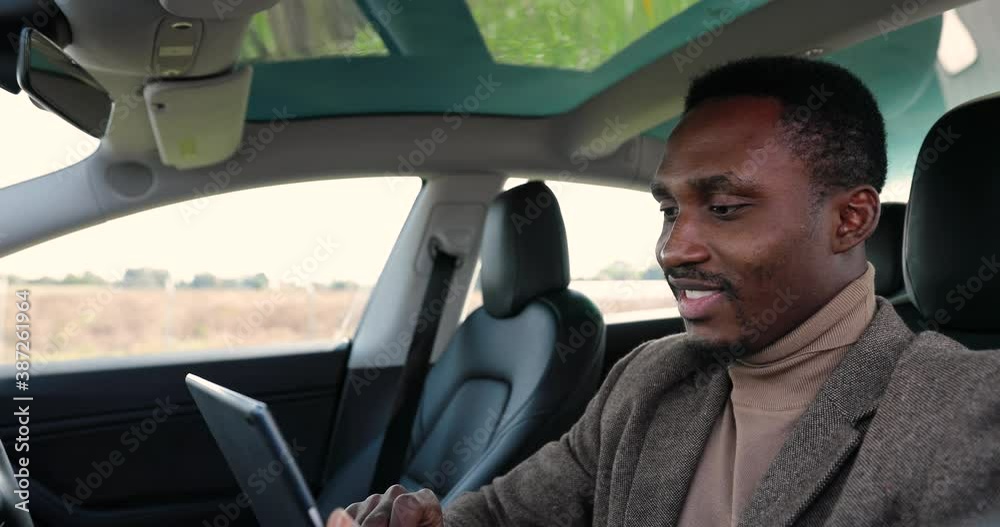 Close up of cheerful handsome African American man in electric automobile smiling and searching internet on tablet. Young happy male sitting in modern car and tapping on device. Business concept Close up of cheerful handsome African American man in electric automobile smiling and searching internet on tablet. Young happy male sitting in modern car and tapping on device. Business concept