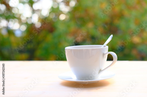 close-up of female hand placing hot ceramic white coffee cup with smoke on saucer over wooden table in nature green background, outdoor morning tea concept, seasonal mood