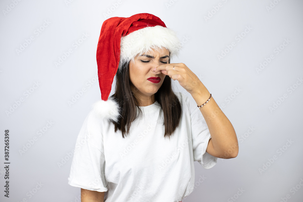 Young beautiful woman wearing a christmas hat over white background smelling something stinky and disgusting, intolerable smell, holding breath with fingers on nose Young beautiful woman wearing a christmas hat over white background smelling something stinky and disgusting, intolerable smell, holding breath with fingers on nose